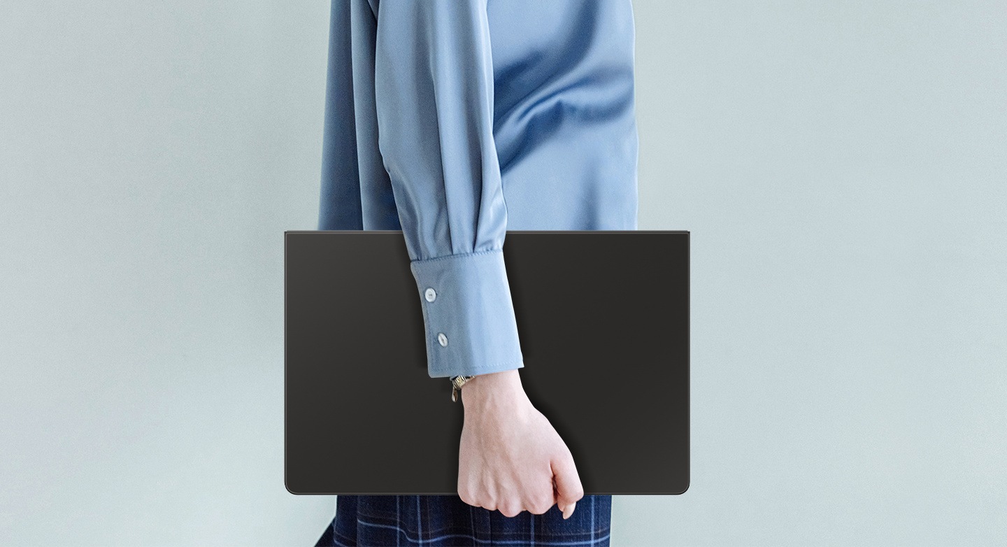 Mavi gömlekli biri elinde Book Cover Keyboard Slim takılı bir Galaxy Tab S10 FE+ ile yürüyor.
