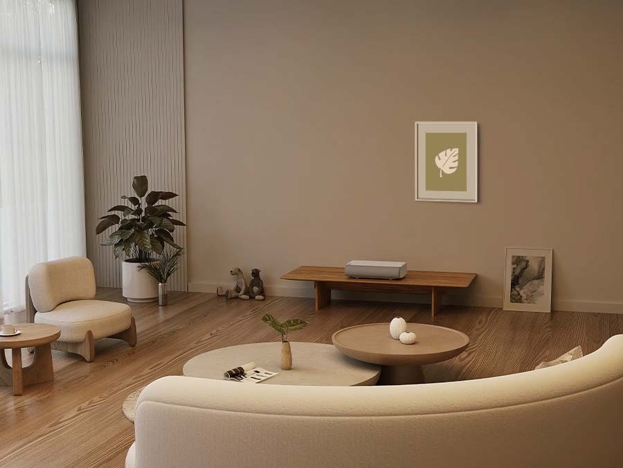 The Premiere projects an ambient screen onto the wall of a living room, over a framed picture of a leaf. The ambient screen depicts a framed ocean picture and a shelf with decor, which blends in with the room. The framed leaf picture  has a dynamic background that changes from palm trees to forest.