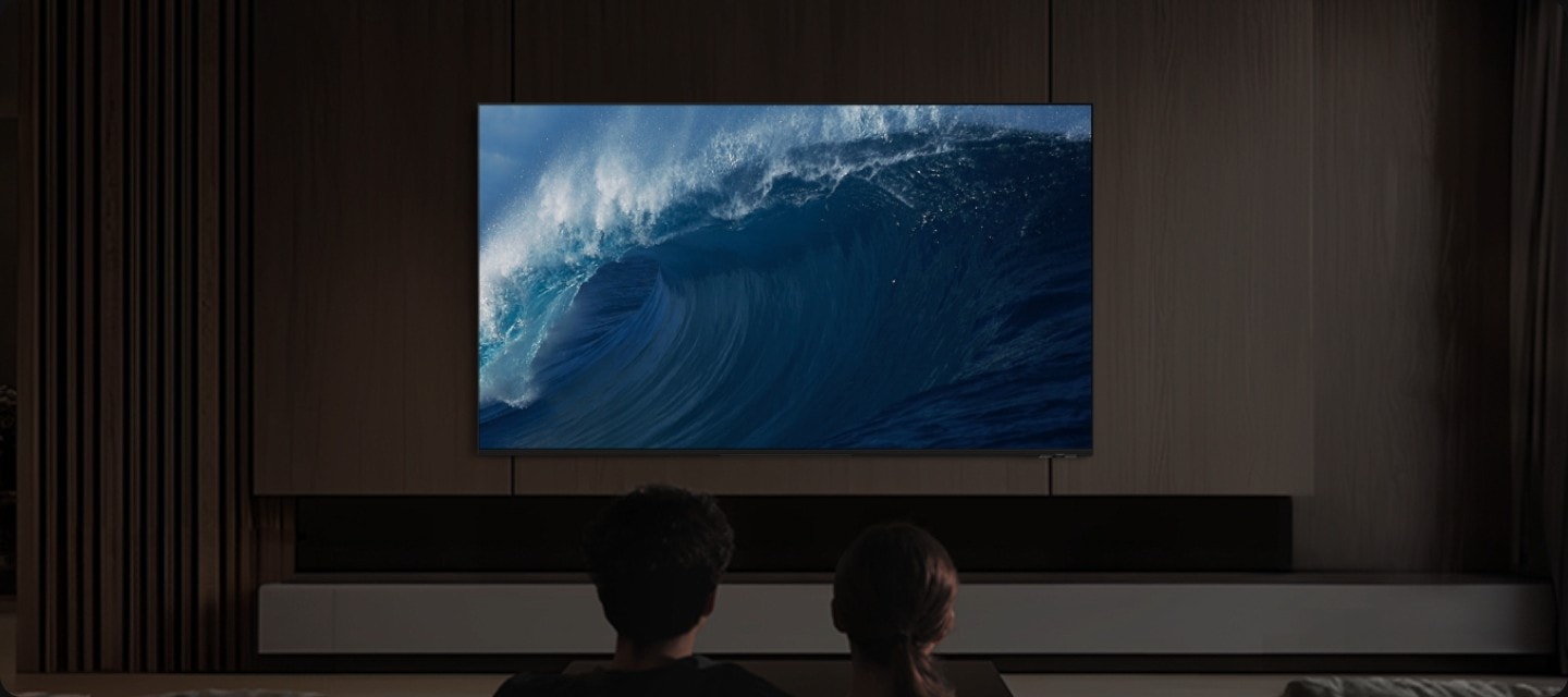 Bir çift, oturma odasında bir koltukta oturuyor ve TV izliyor. TV’nin ön hoparlörleri aydınlanıyor ve TV’nin yan tarafından ses dalgaları yükseliyor. Birden fazla yönde yukarıya doğru ses veren hoparlörler ise çifti bir ses kubbesi altında çevreleyerek odayı dolduruyor. Dolby Atmos logosu.