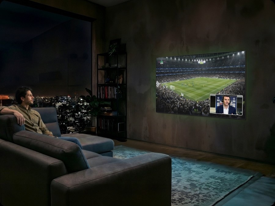 Bir adam gece Samsung TV’de futbol maçı izliyor. TV’nin yanından ve önünden ses dalgaları yayılıyor. Uzaktan kumandaya konuşarak “Futbol spikerinin sesini yükselt” diyor. AI Football Mode Pro, spikerin sesini otomatik olarak artırıyor ve bu işlev, TV’nin önünde büyüyen ses dalgalarıyla gösteriliyor.