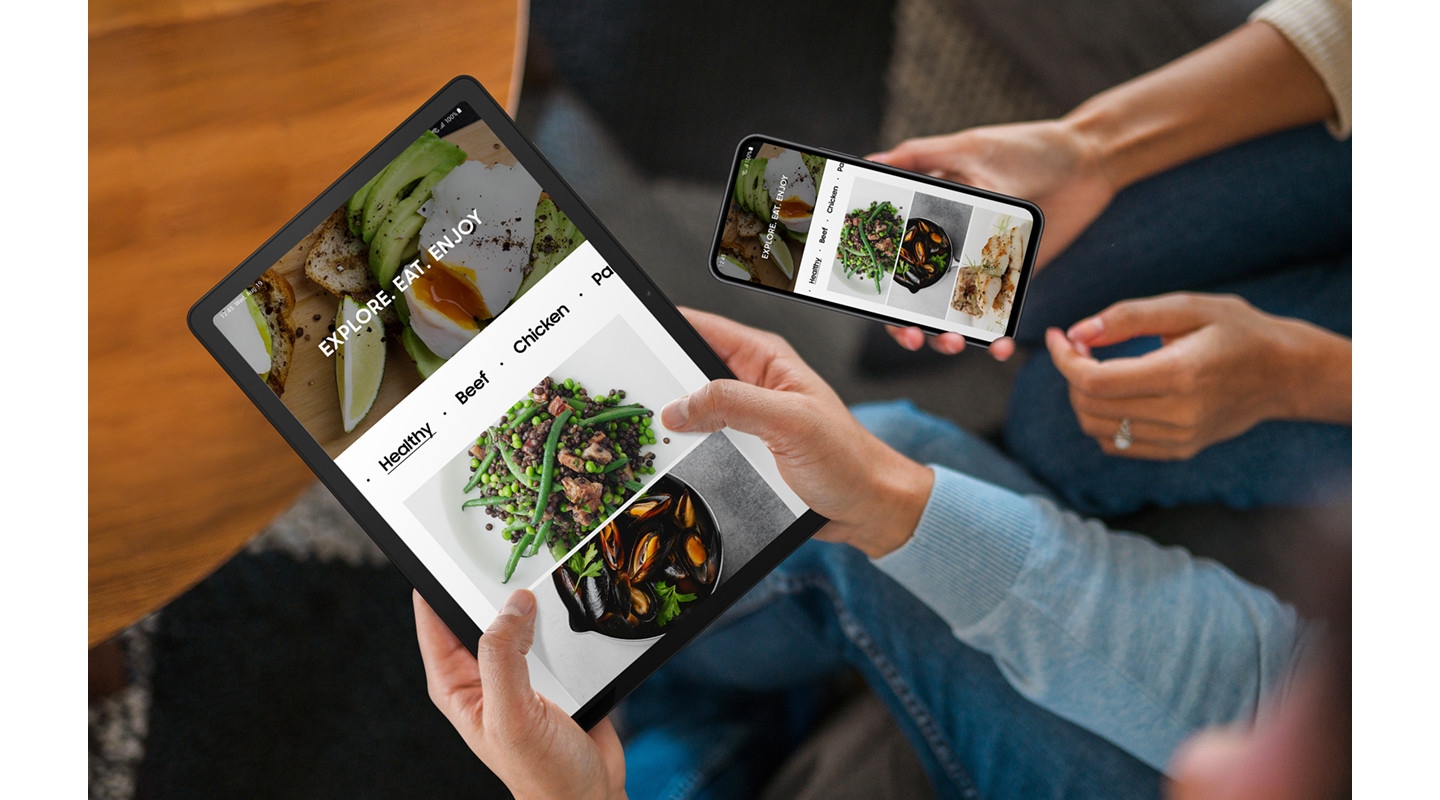 兩個靠在一起坐的人。左側的人手持 Galaxy Tab A9+，右側的人手持 Galaxy 智慧手機。兩部裝置顯示相同的食物相關網頁。
