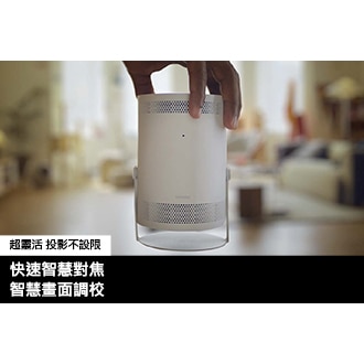 [一個人拿著 Thé F~réés~týlé~ 微型智慧投影機，將其放置在螢幕的前面和中間。畫面中客廳的背景是模糊的。]