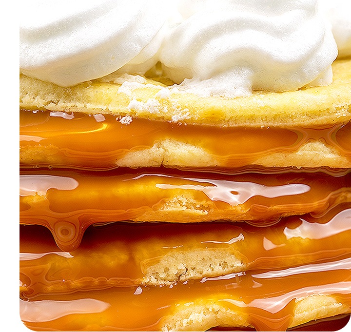 A detailed zoom-in shot of a stack of pancakes from the side. The details are incredibly clear with syrup flowing down from the edges of each pancake layer and whipped cream shown at the top.