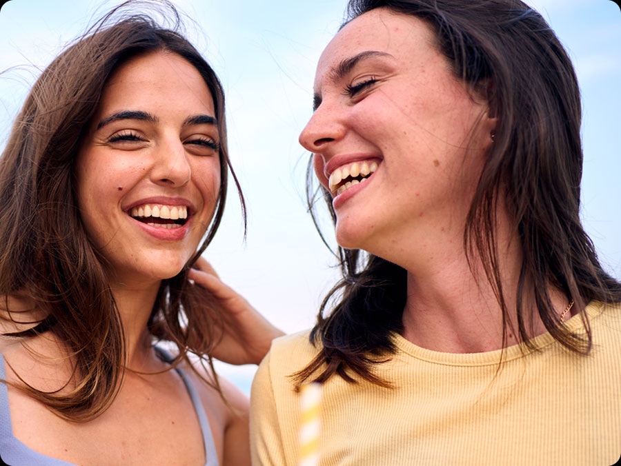 Two people stand close together outdoors. One wears a light blue sleeveless top, the other a yellow short-sleeved shirt. Highlights the Super HDR functional aspect of Galaxy S25 FE.