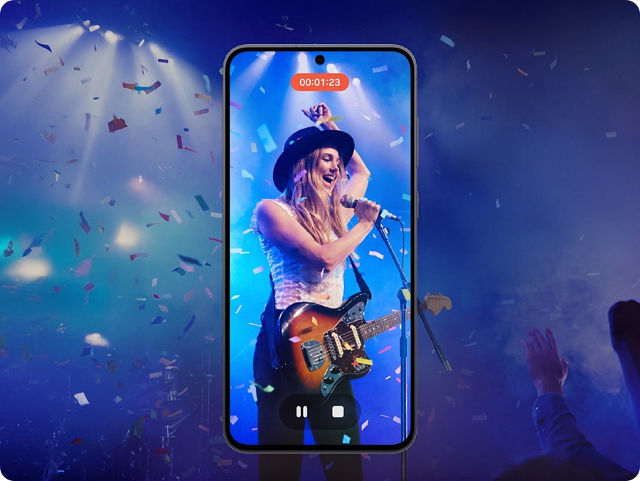 A smartphone screen shows a video recording of a live stage performance of a performer playing an electric guitar. The smartphone screen shows that the video recording is vibrant to highlight that clear videos can be taken even in dark settings.