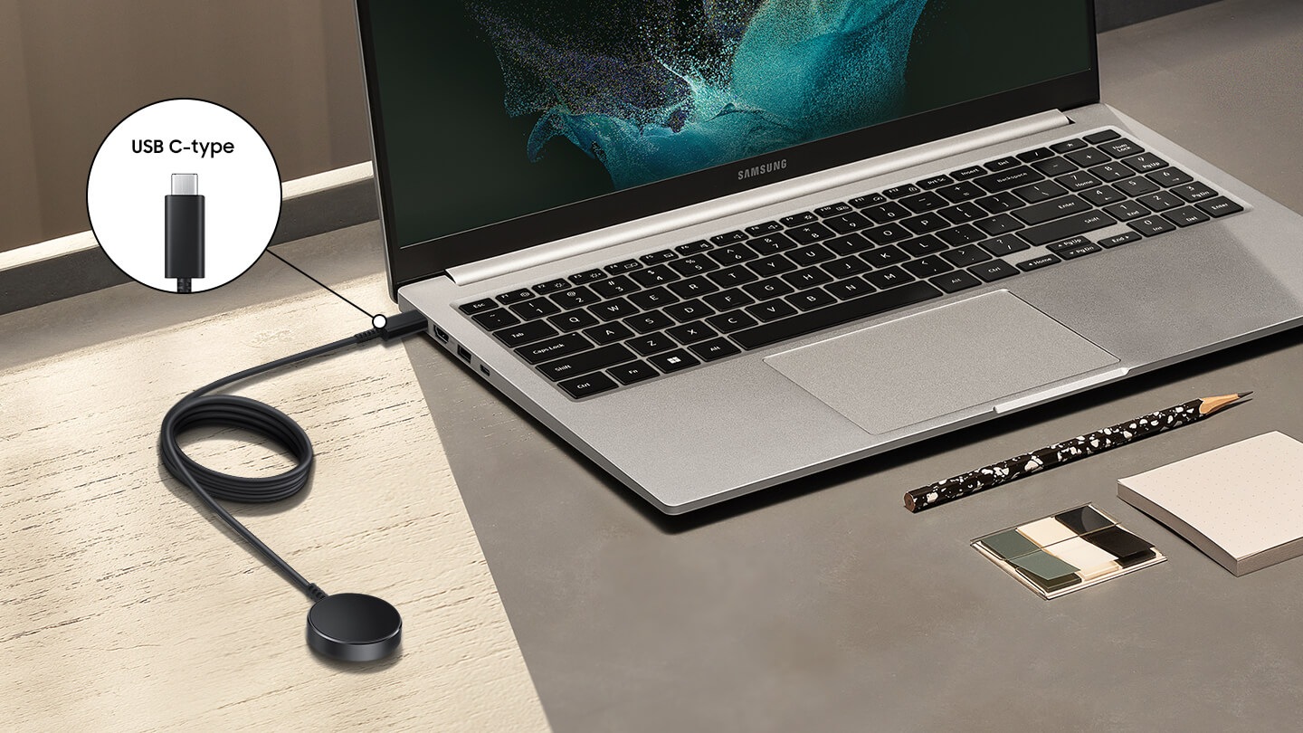 An angled view of a Samsung laptop open on a desk with the charging cable plugged into a USB-C port.