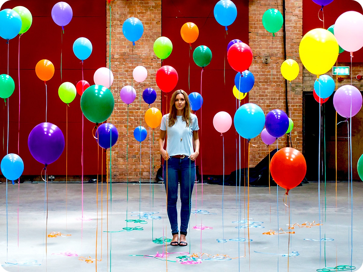 1. There is a close-up shot of a woman standing in the middle of many different color balloons.