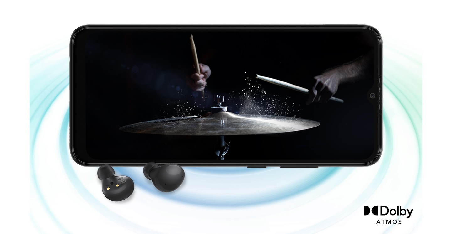 Galaxy A03 in landscape mode and an image with a person playing drums in the black background onscreen. A pair of black Galaxy Buds2 are placed in front of the device. On the right bottom is a logo for Dolby Atmos.