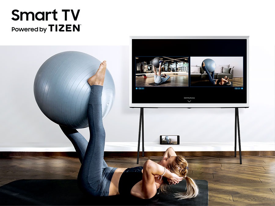 A woman is working out with an exercise ball on the ground with her smartphone next to her. The Frame is displaying what's on her smartphone as well as a video of a trainer on the TV's screen.