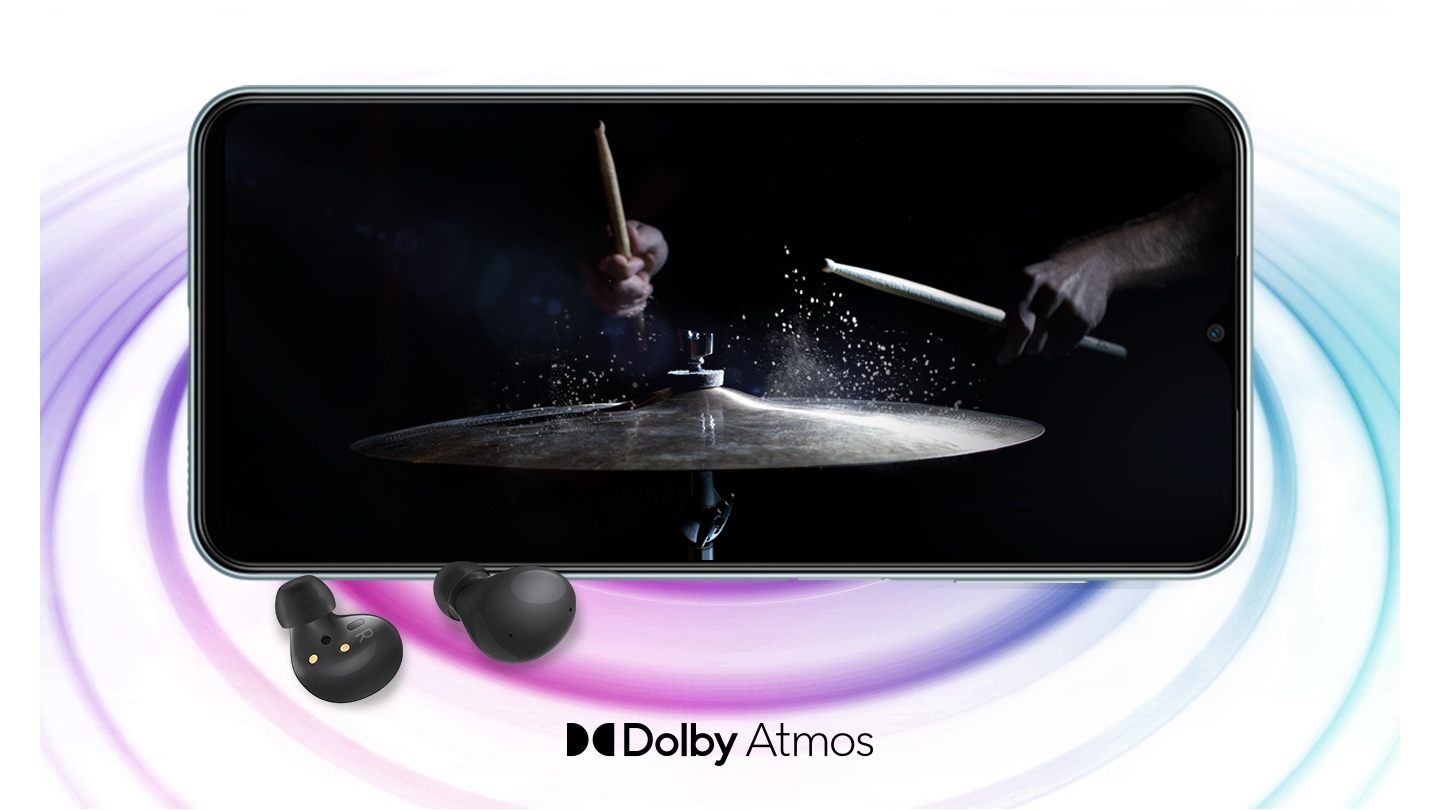Galaxy M23 5G in landscape mode and an image with a person playing drums in the black background onscreen. A pair of black Galaxy Buds2 are placed in front of the device. On the right bottom is a logo for Dolby Atmos.