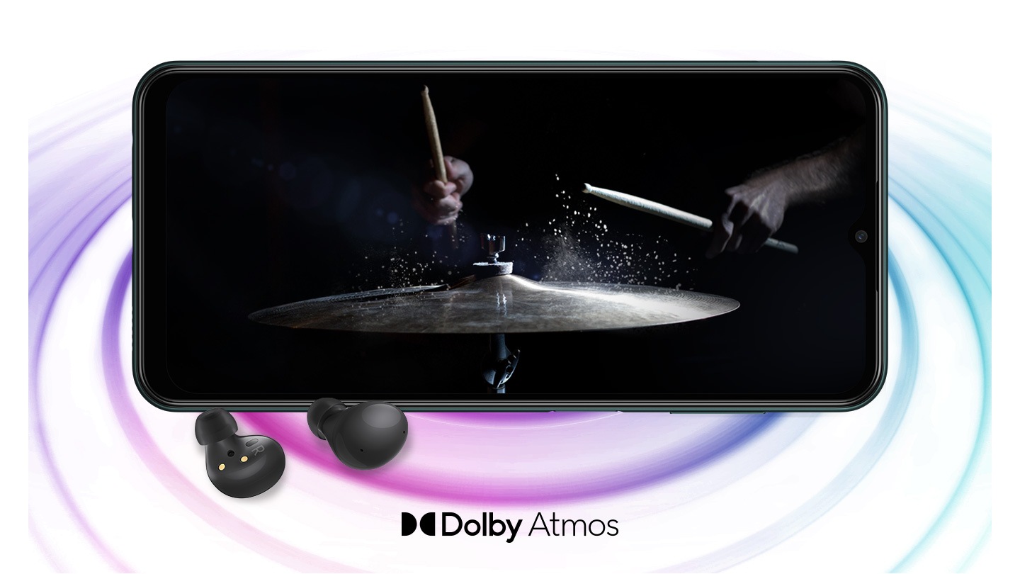 Galaxy M13 în modul peisaj și o imagine cu o persoană cântând la tobe pe fundalul negru de pe ecran. O pereche de Galaxy Buds2 negre sunt plasate în fața dispozitivului. În partea de jos este un logo pentru Dolby Atmos.
