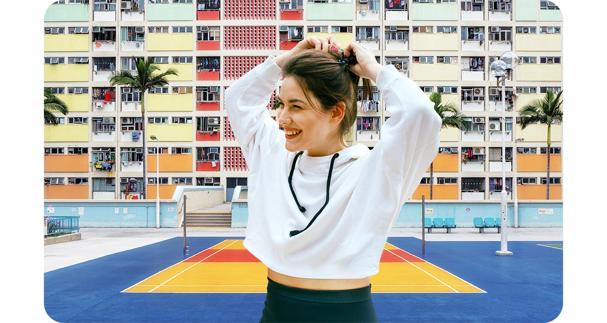 Portrait mode is off. A woman is fixing her hair and smiling, looking to her right. Behind her is a is volleyball court, colorfully painted apartments, and tropical trees.