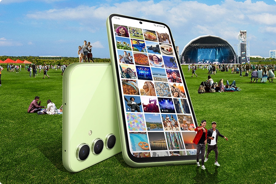 In an outdoor concert hall setting, many people are sitting on the grass. In the foreground, two large Galaxy A54 5Gs in Awesome Lime are also sitting on the grass, overlapping to show both the backside and the front screen. On the screen, the Gallery app is opened to show many pictures in its album.