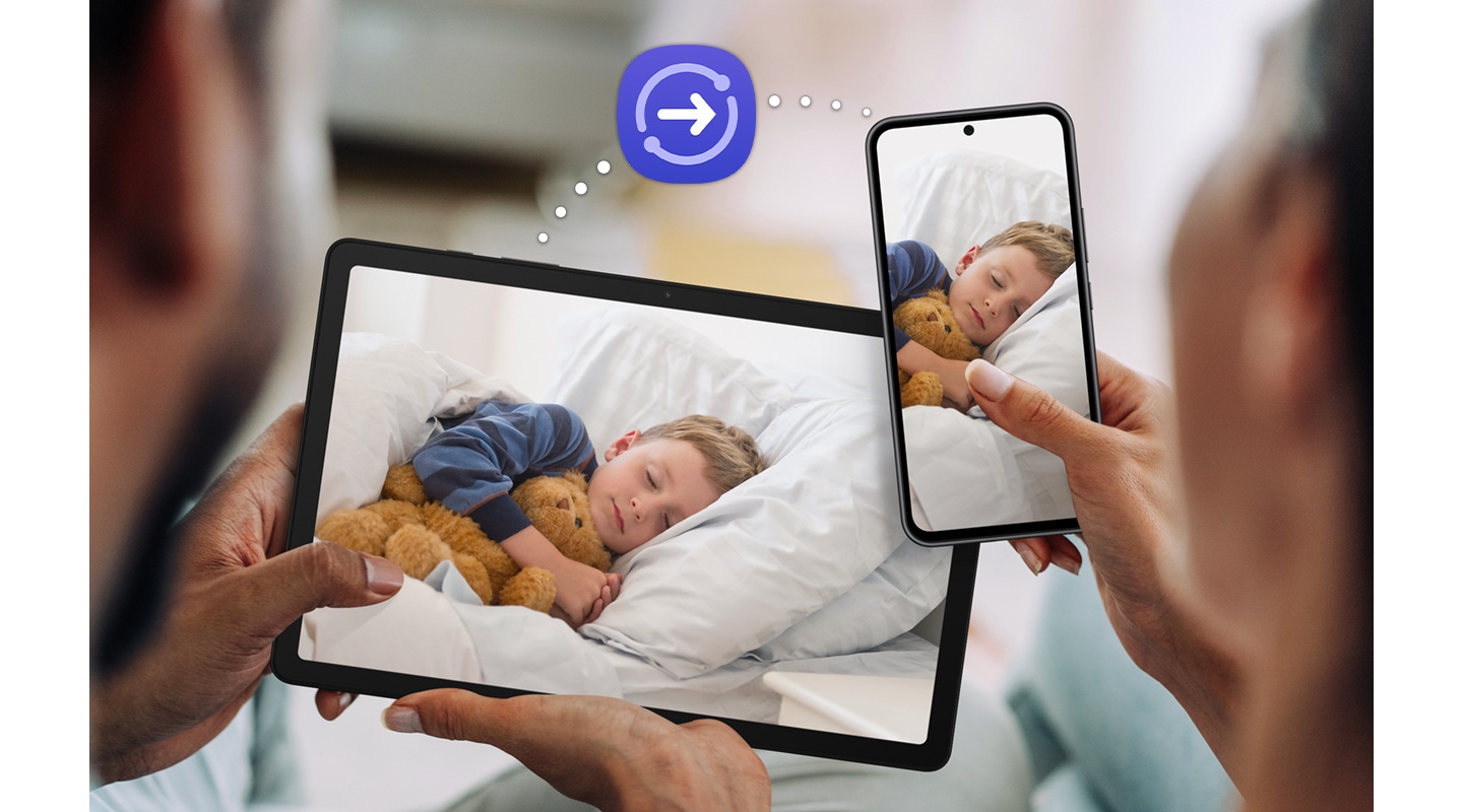 A couple is holding up Galaxy Tab A9+ and a Galaxy smartphone, both with the same photograph of a sleeping child onscreen. Above the devices is a dotted line that connects the devices, with the Quick Share icon in the middle to indicate the file-sharing feature.