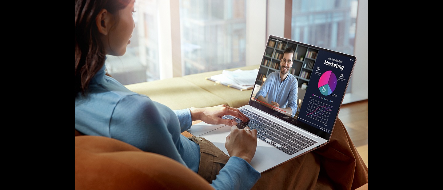 A woman is sitting on a couch in a bright room with Galaxy Book4 Pro on her lap A video played onscreen is clear and vibrant with improved visibility thanks to Vision Booster