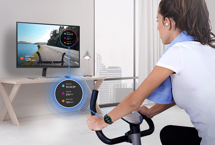A woman is riding an indoor bicycle. She is wearing a Galaxy Watch and Buds, and her watch screen is overlaid on the monitor.
