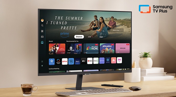 There is a monitor on a desk and its home UI is shown with OTT applications. On the upper right, there's a logo of Samsung TV Plus.