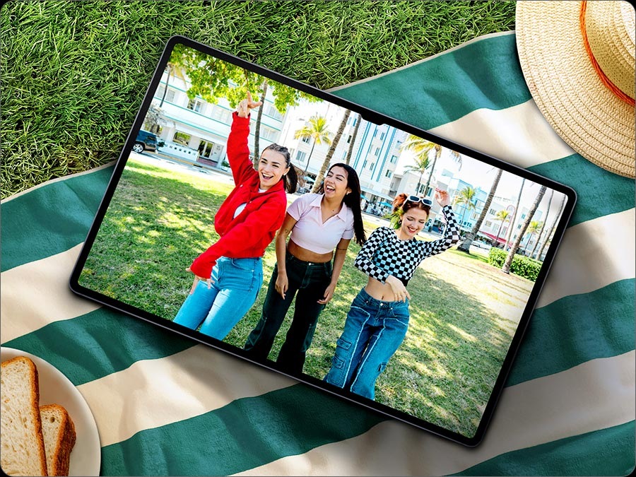 A Galaxy Tab S10 Series device, placed on a picnic blanket outdoors, displaying a video recording of three women posing happily. The screen is clear with minimal reflection, thanks to anti-reflection coating.