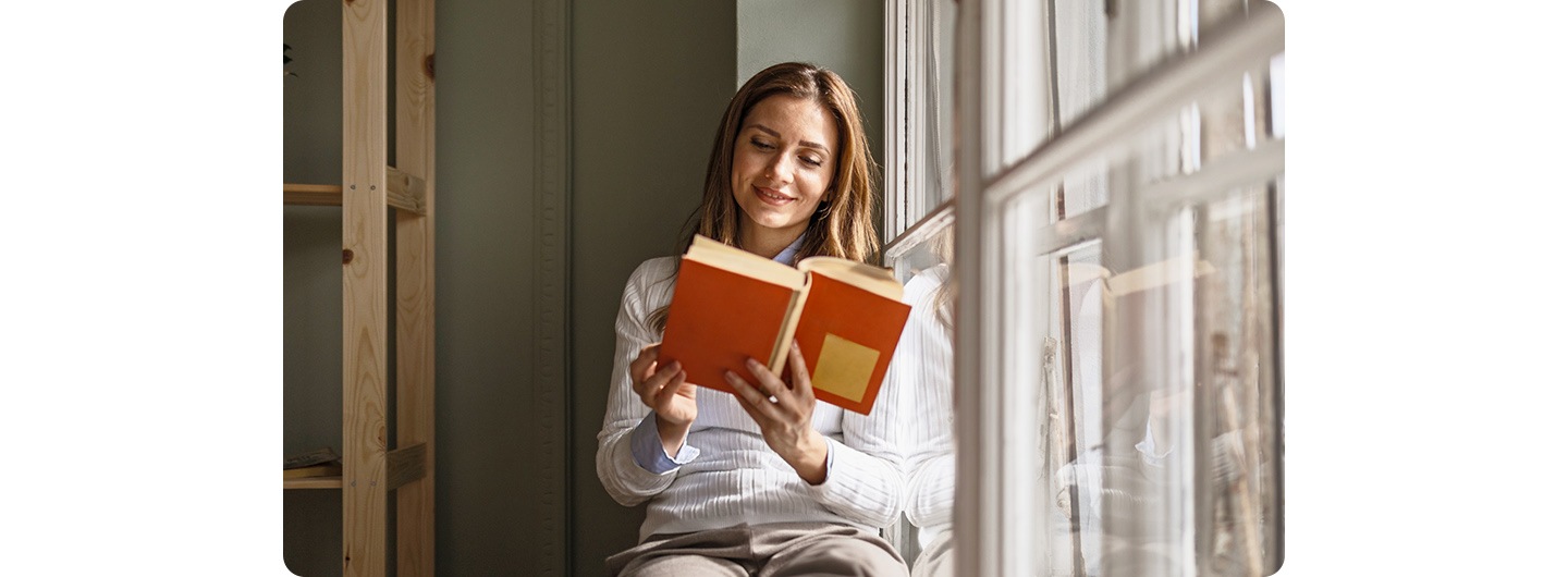 A woman sitting indoors, reading by a window. A line down the screen splits it into two sections, comparing a low-light shot on the left with a bright-light shot on the right. 