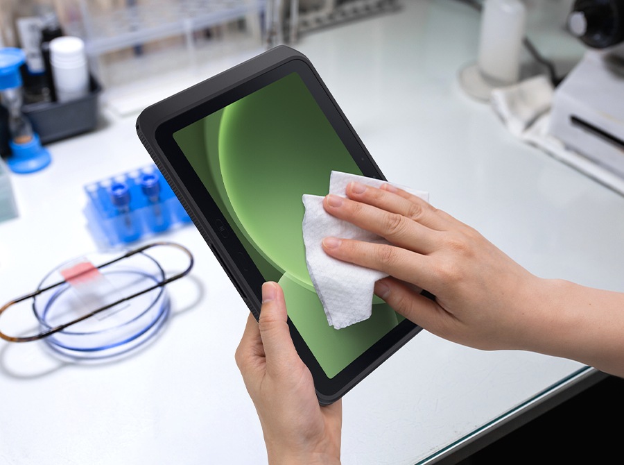 A hand holds Galaxy Tab Active5 Pro while the other wipes the screen with a cloth in a lab setting, illustrating that the device can be cleaned with sanitizer. A green-shaded shape is visible onscreen.