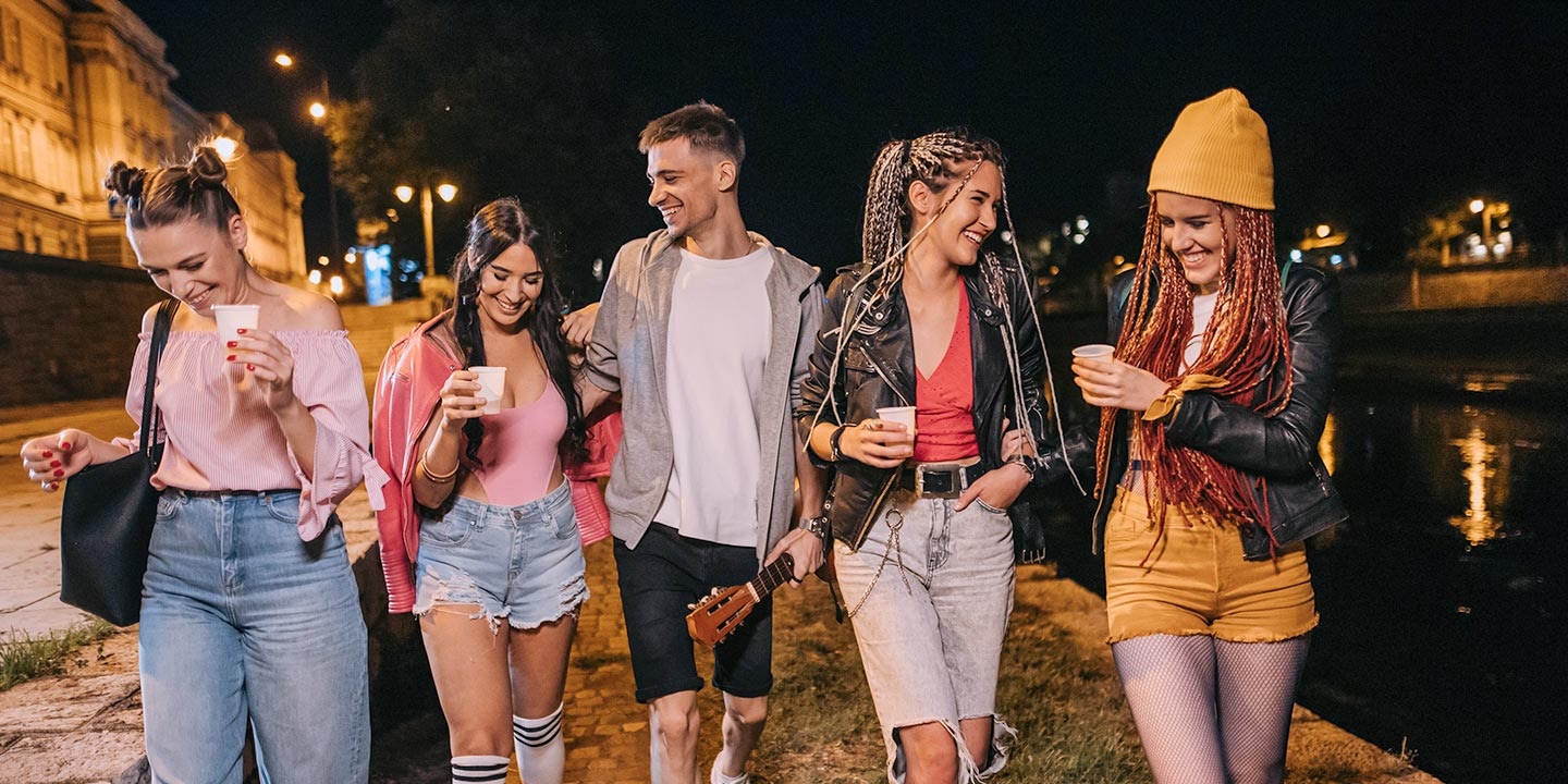 Five friends, four women and one man, walk beside a river at night holding drinks under streetlights. A vertical slider splits the image, with the left side dim and muted and the right side bright and colorful, revealing clearer details like a yellow beanie, ripped jeans, and reflections on the water.