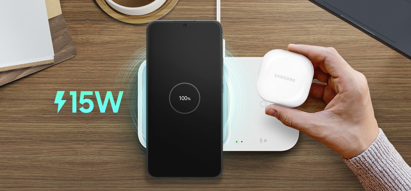 Device in centre view being placed on the wireless charger on top of a wooden surface with a blue 15W symbol to the left of the image and a hand holding another device ready to be charged on the right of the device