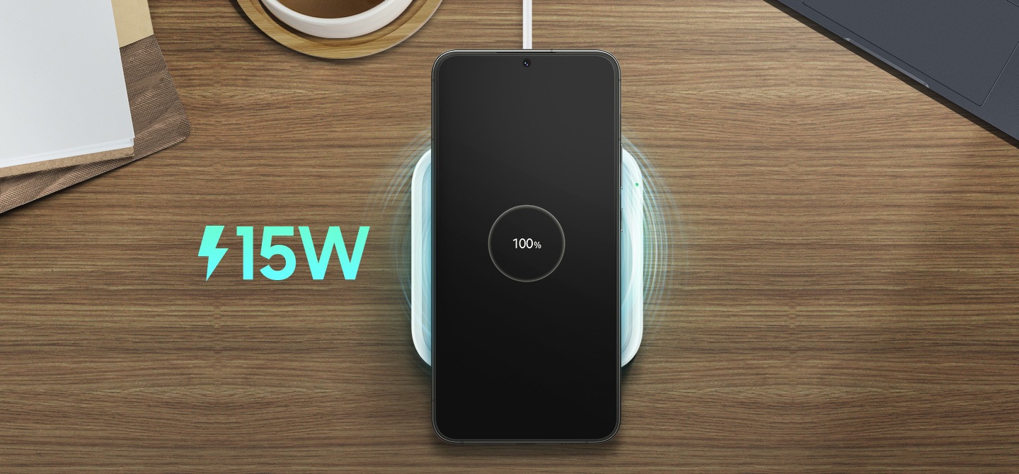 Device in centre view being placed on the wireless charger on top of a wooden surface with a blue 15W symbol to the left of the image and a hand holding another device ready to be charged on the right of the device.