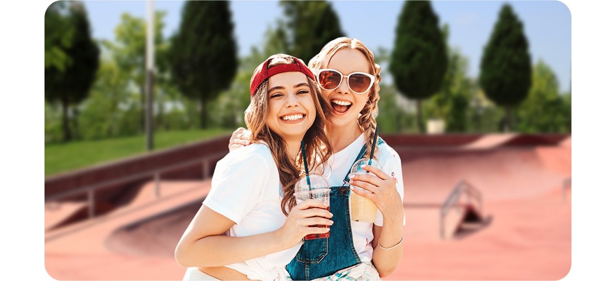 live focus on activated, Two female friends are smiling, looking into the camera in front of a skatepark with trees and grass in the background. With the Live Focus On icon above the shot activated, the background is blurry.