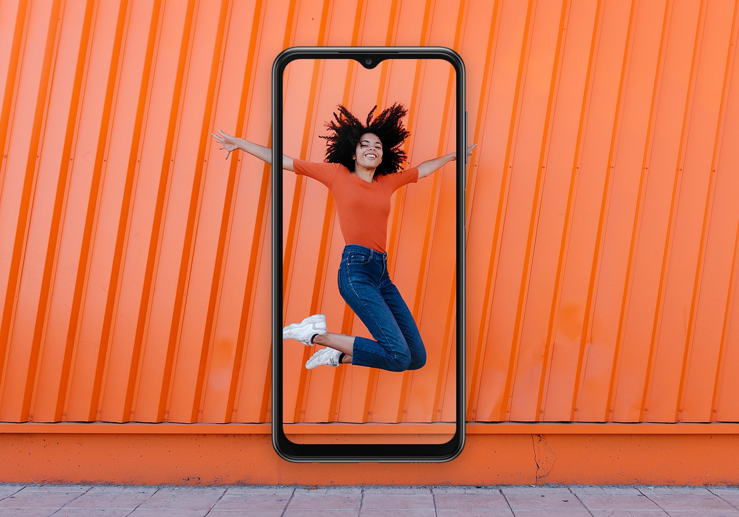 A woman, smiling, is in mid-jump in front of an orange wall background. At the center, a Galaxy A23 is overlapping and captures the woman inside the screen.