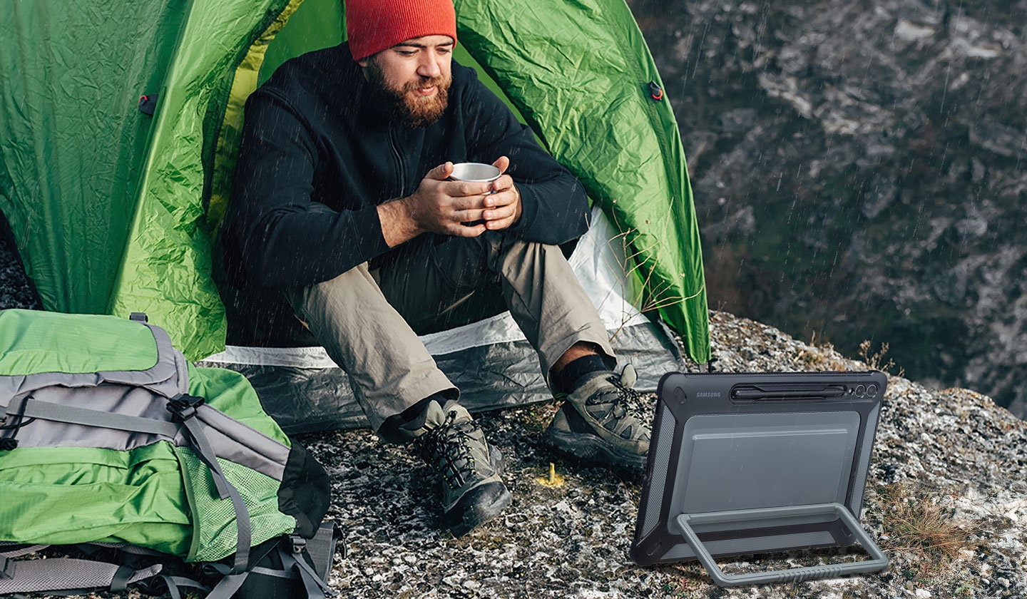 Một người đàn ông cầm cốc đang ngồi trước lều cắm trại trên bề mặt đá, nhìn vào thiết bị Galaxy Tab S9+ có Ốp lưng siêu bảo vệ, được đặt theo chiều ngang bằng chân chống ở phía sau.
