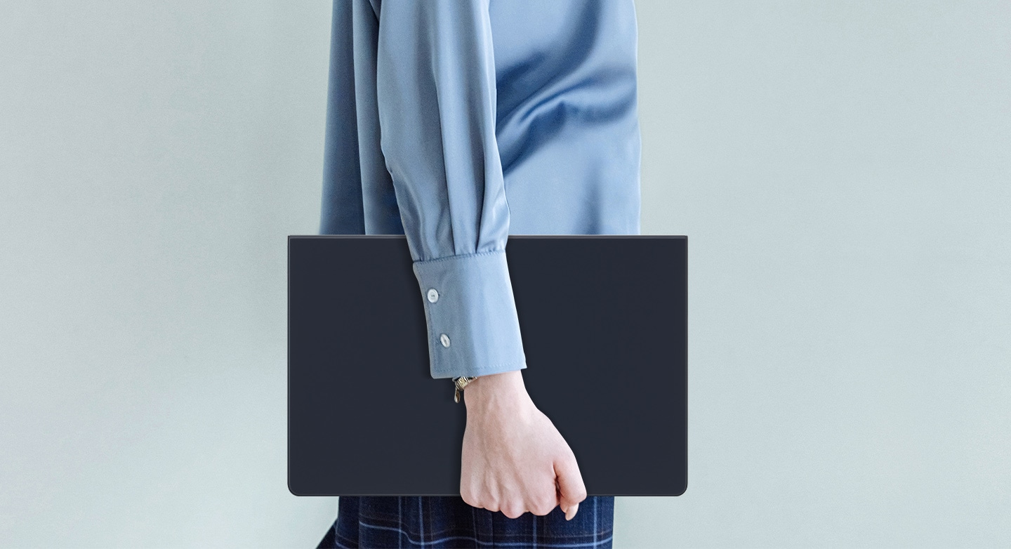 Một người mặc áo sơ mi màu xanh dương đang đi bộ trong khi cầm một chiếc Galaxy Tab S9+ đang sử dụng Book Cover Keyboard Slim.