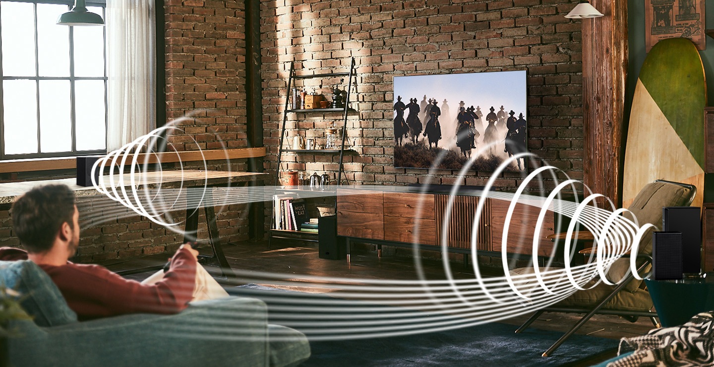 A man enjoys racing content on his TV. Soundwave graphics are playing from Samsung Wireless Rear Speaker Kit and Soundbar, demonstrating Wireless Surround Sound Compatible feature of Samsung soundbar.