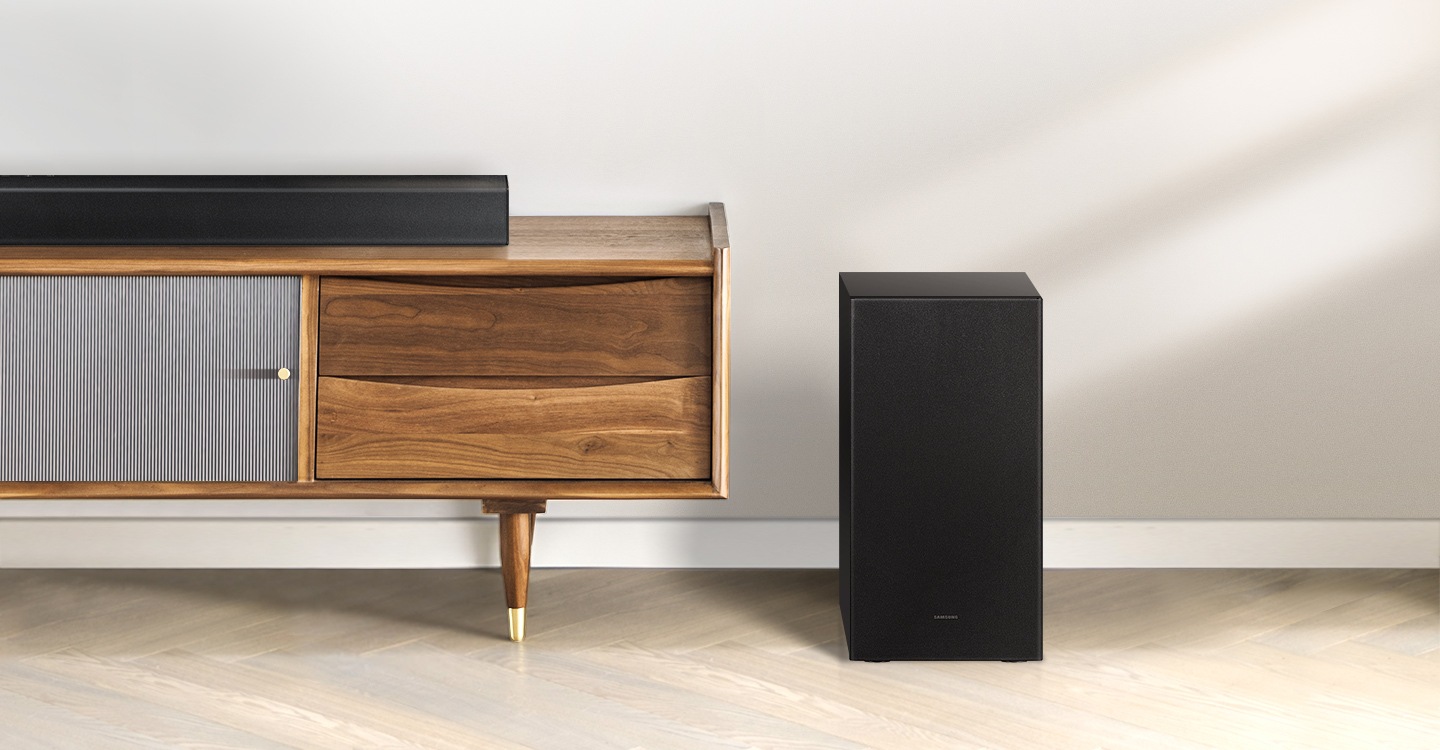 Samsung A series soundbar is being shown on top of a contemporary TV cabinet along with its matching subwoofer which is being displayed to the side of the TV cabinet.