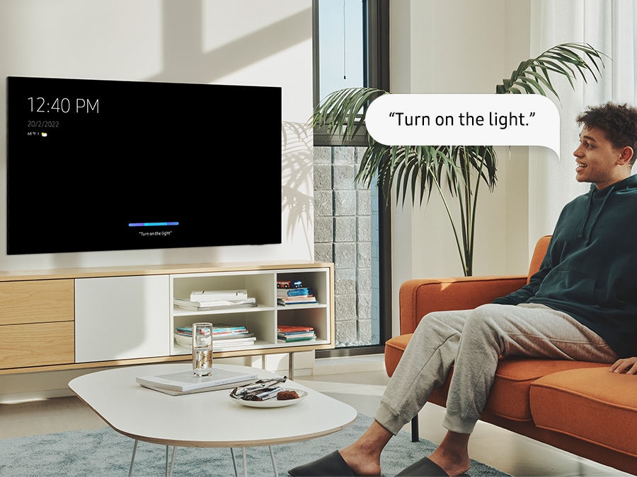A man is saying "Turn on the light." to a QLED TV and the TV is responding to the command with the Always On Voice feature.