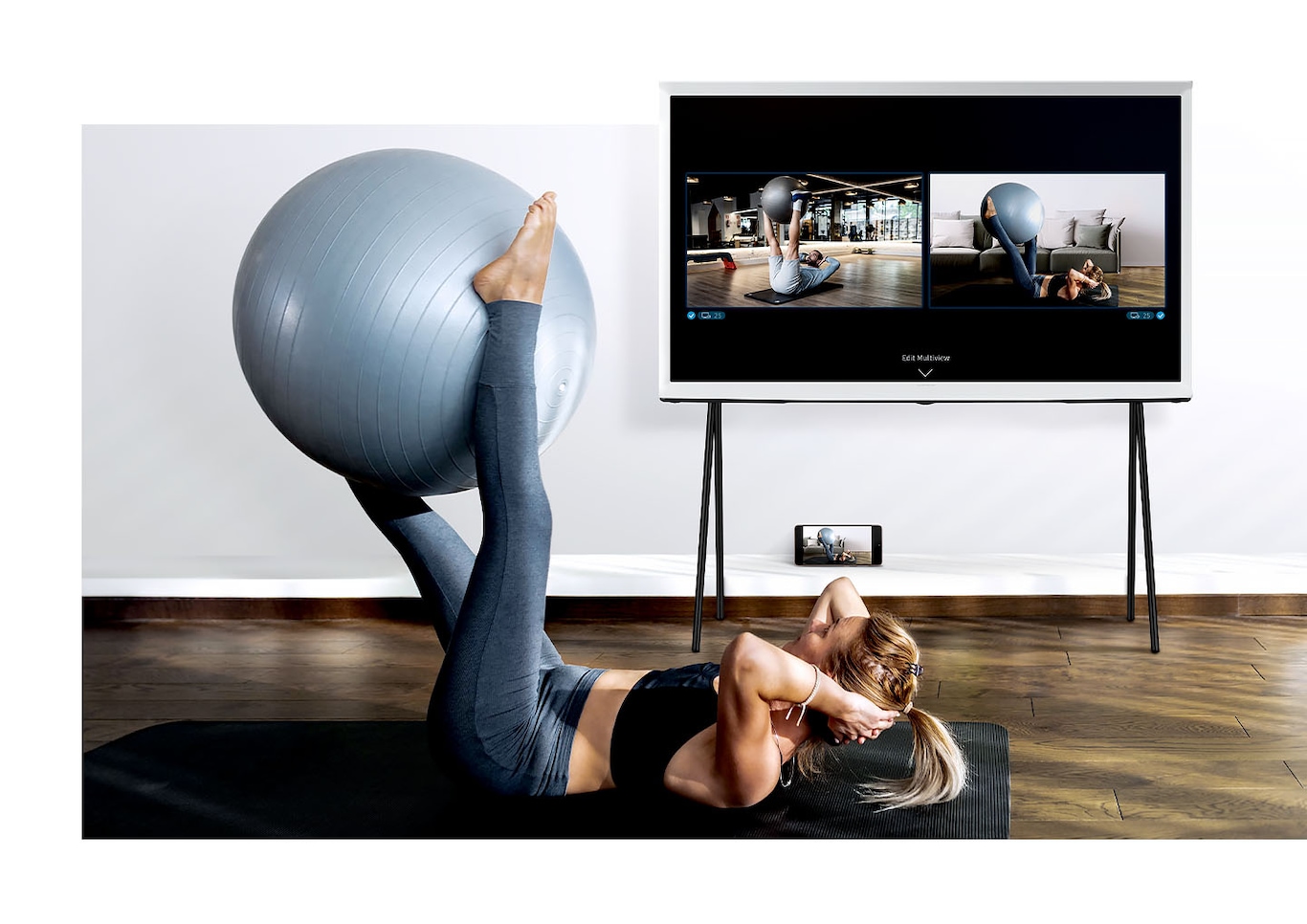 A woman is working out with The Serif's Multi View function to see her reflection and a trainer's demonstration side by side.