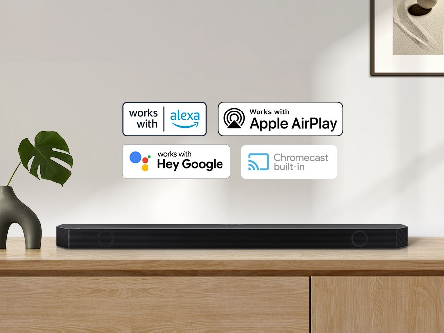 Alexa logo, Apple AirPlay logo, Hey Google logo, and Chromecast Built-in logo can be seen along with Samsung Q990B soundbar which is sitting on living room cabinet.