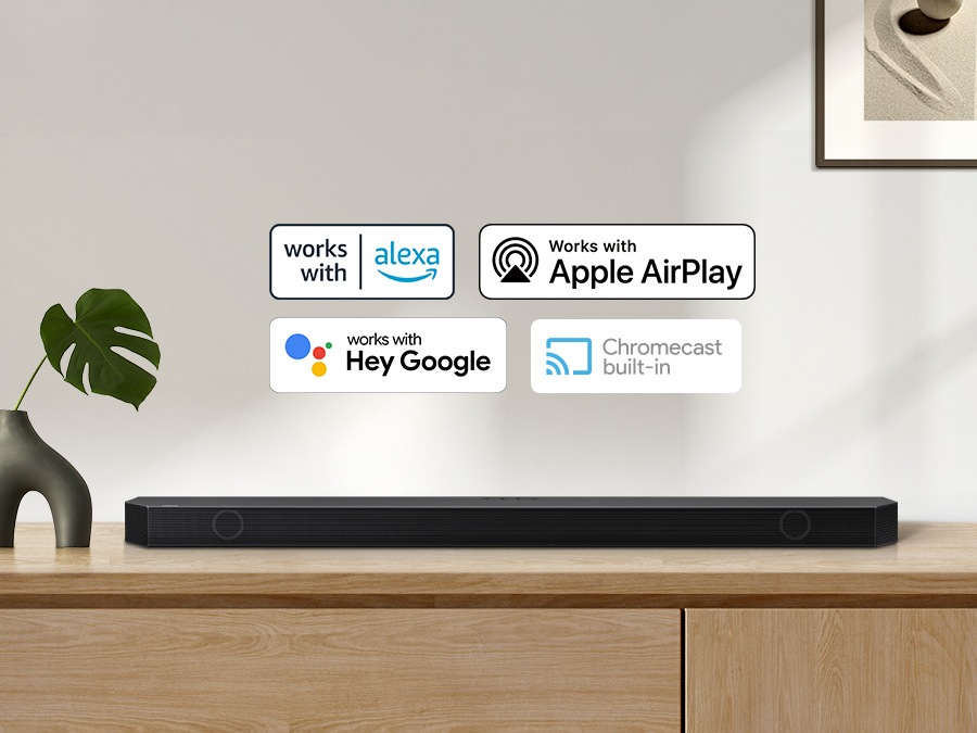 Alexa logo, Apple AirPlay logo, Hey Google logo, and Chromecast Built-in logo can be seen along with Samsung Q930B soundbar which is sitting on living room cabinet.