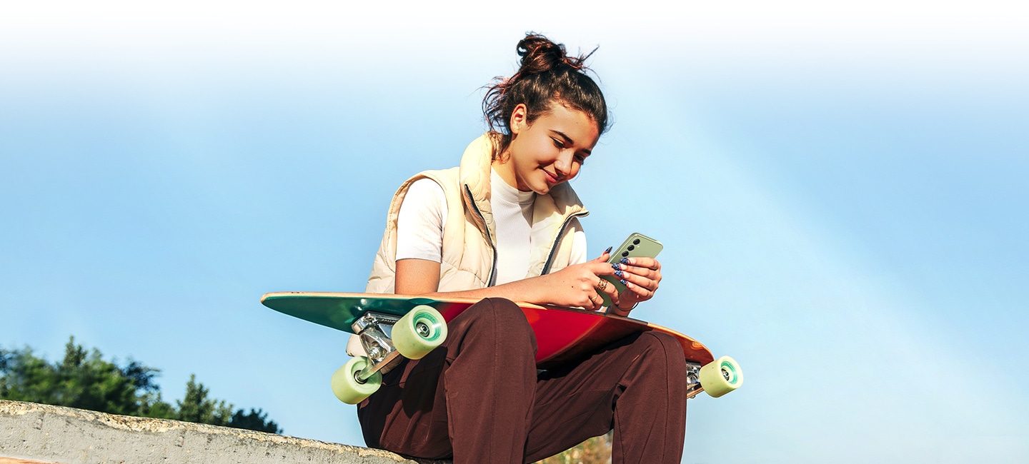 A woman is looking at Galaxy A54 5G under bright sunlight, with a skateboard on her lap. 