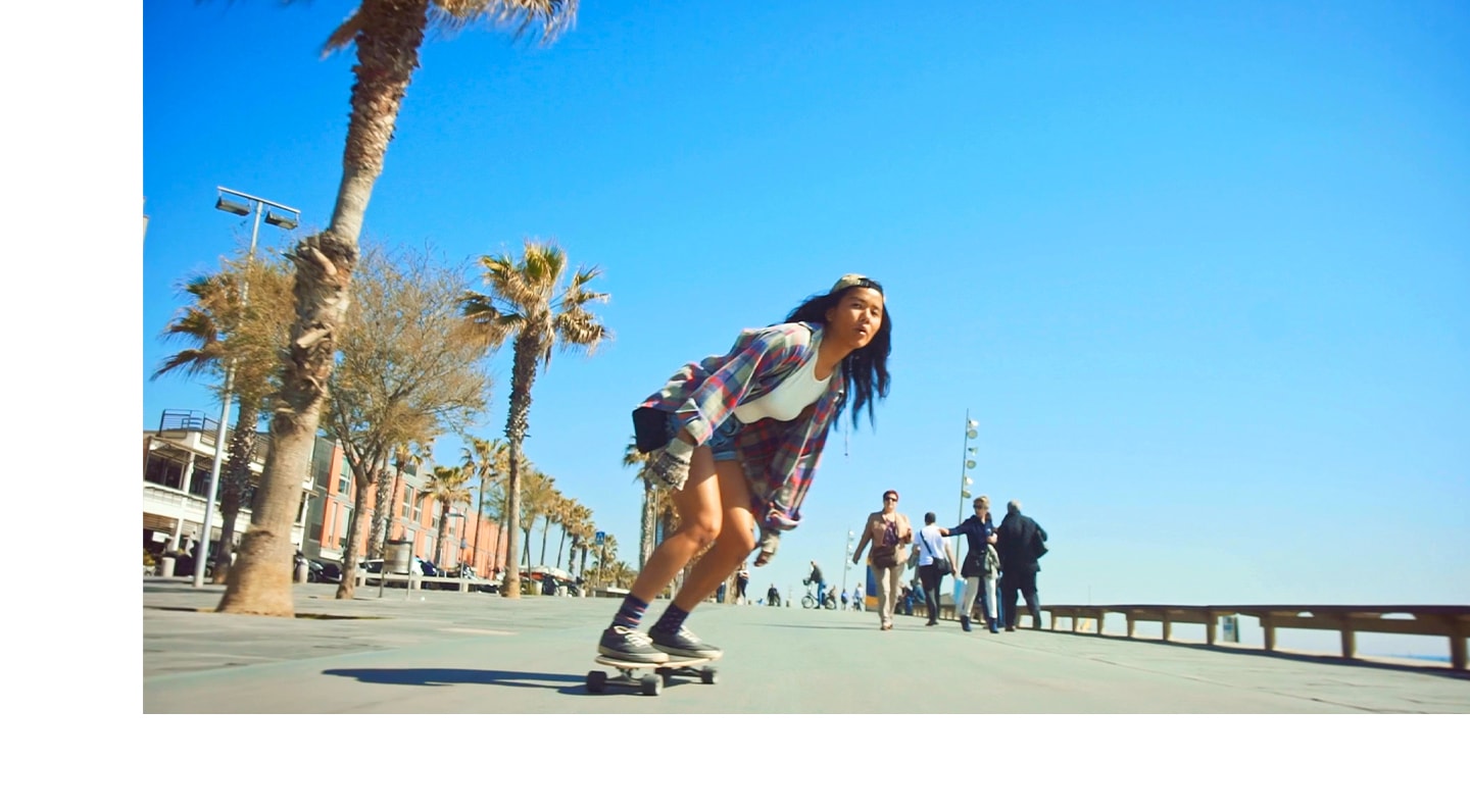 A girl on a skateboard is skating in the middle of a street with a pier to her left and palm trees to her right. As the girl zigzags on her skateboard, the camera captures vivid details and stabilized images of the people sitting at the pier, people walking and riding bicycles.