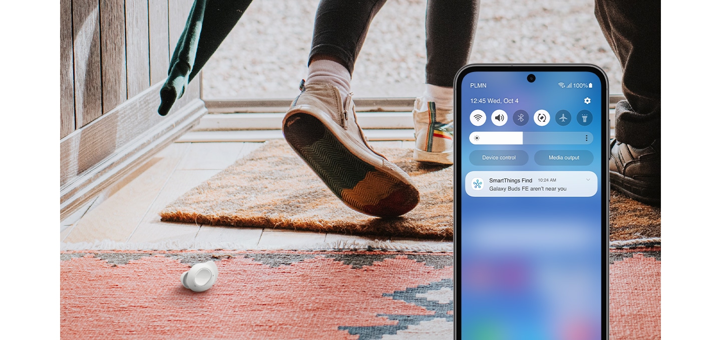 A smartphone display with the app SmartThings Find on. A person is seen walking out of a location and one Galaxy Buds FE earbud is left on the carpeted floor. The device's screen shows a notification message that reads Galaxy Buds FE aren't near you.