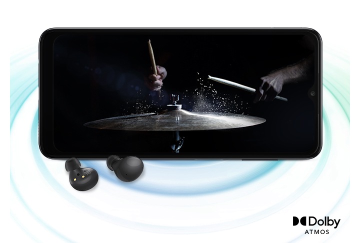 Galaxy A04s in landscape mode and an image with a person playing drums in the black background onscreen. A pair of black Galaxy Buds2 are placed in front of the device. On the right bottom is a logo for Dolby Atmos.