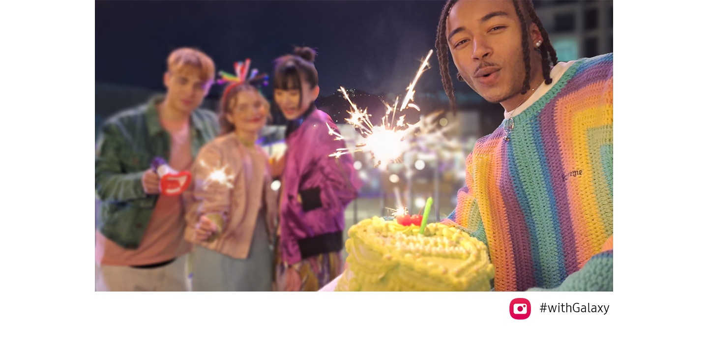 Three friends are on top of a bridge at night, celebrating with sparklers. When another friend enters the frame from the right, holding a cake, the Auto-framing feature automatically adjusts the frame to include all of them as well as the focus on the friend holding the cake, who is standing closer