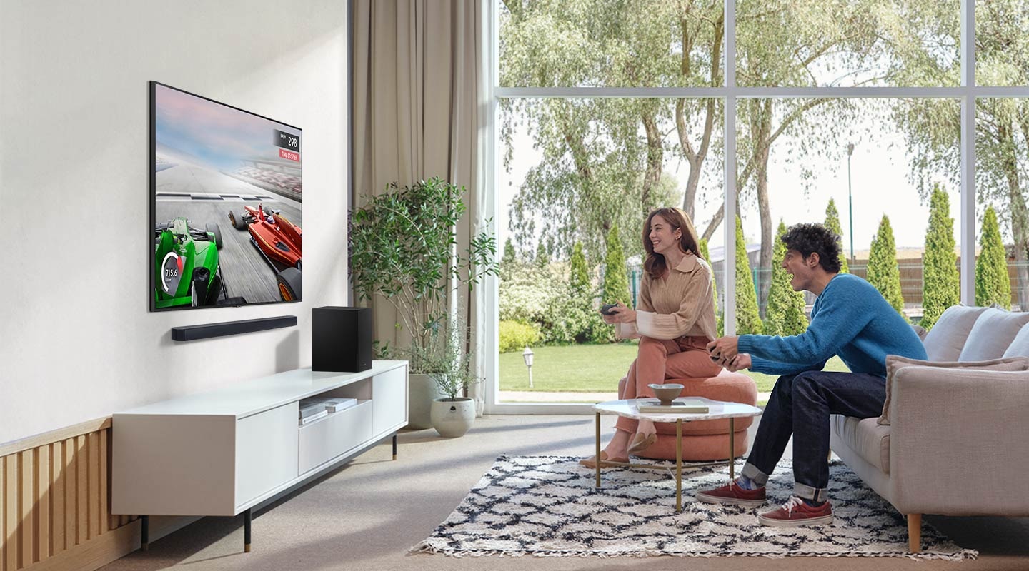 A man and woman enjoy the Soundbar’s Game Mode while playing a racing game on their TV connected to Soundbar and subwoofer.