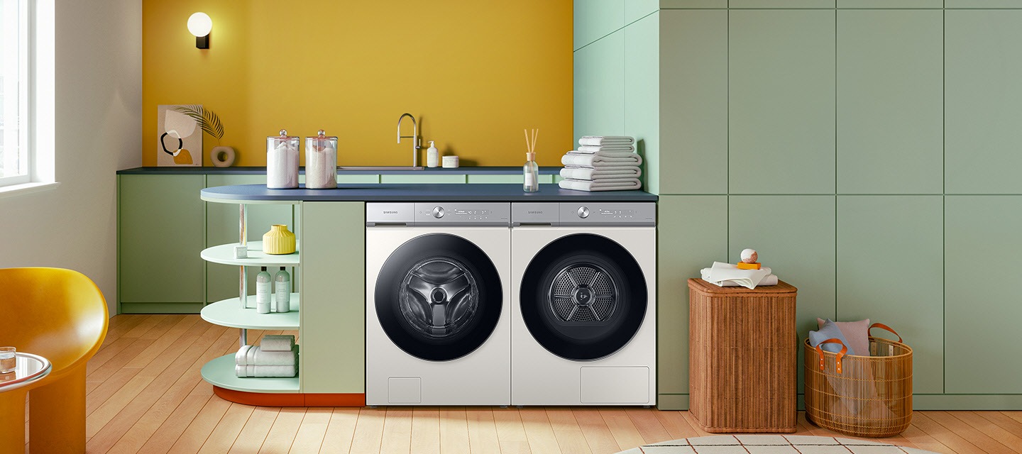 A white Bespoke Grande AI washer and dryer set are built side by side into a green cabinet under a countertop. Towels, detergent, and home accessories are arranged nearby.