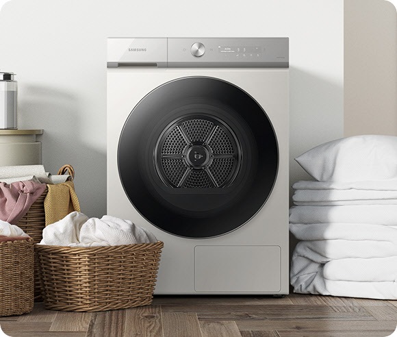 A white Bespoke Grande AI dryer is in a laundry room. Three baskets of laundry are placed to the left of the machine, and folded white bedding is placed to the right of the machine.