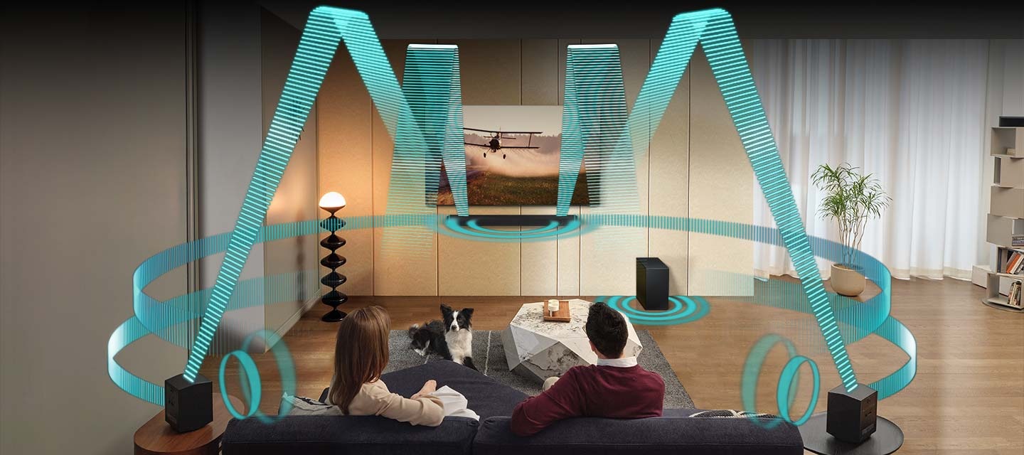 In a living room, a couple sits on a sofa and watches TV. The TV is installed onto the wall, with a Soundbar directly underneath. A pair of rear speakers are set on side tables to the back of the room. Sound waves emanate from the TV, Soundbar and rear speakers from various angles, traveling in multiple directions to fill the room.