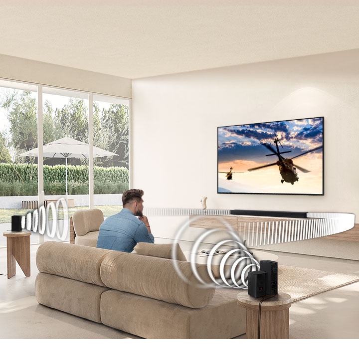 A man watches TV. A Soundbar and rear speakers give off sound waves of different types traveling toward the man.