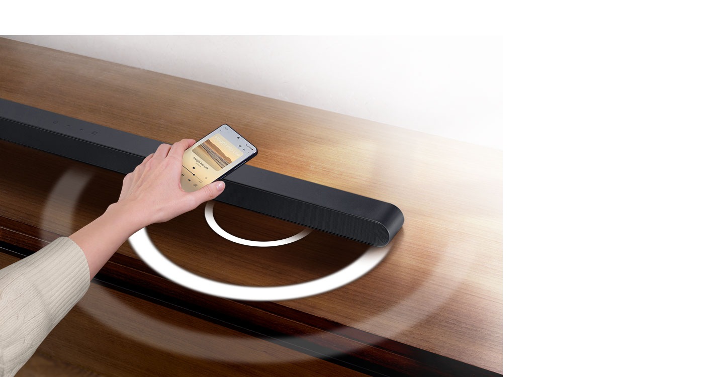 A hand holds a phone playing music and taps it lightly against an Ultra Slim Soundbar. Instantly, the Soundbar gives off a series of round sound waves, indicating that it's playing the same music as the phone.