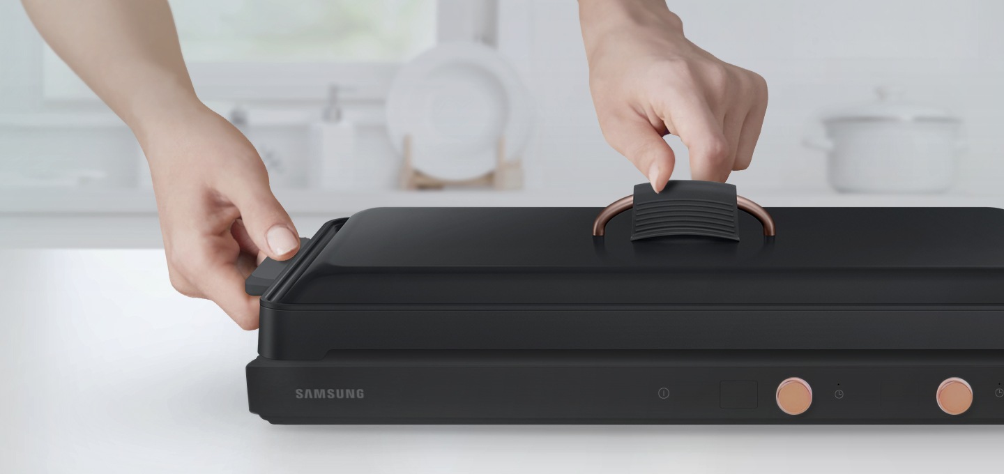 테이블위에 놓인 전용용기에 좌,우측 실리콘 손잡이와 뚜껑 실리콘 손잡이가 결합되어 있고 손으로 뚜껑을 잡는 모습이 보여집니다.