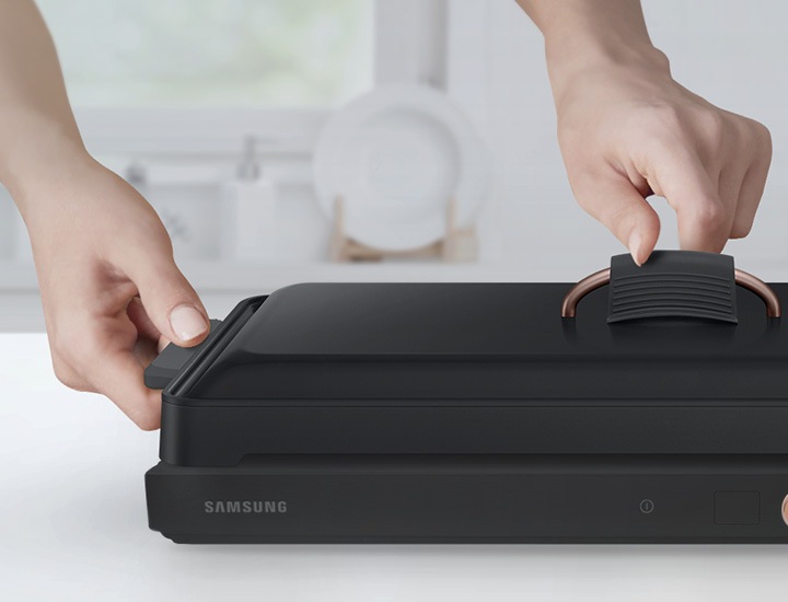 테이블위에 놓인 전용용기에 좌,우측 실리콘 손잡이와 뚜껑 실리콘 손잡이가 결합되어 있고 손으로 뚜껑을 잡는 모습이 보여집니다.